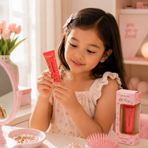 A young girl with dark hair is shown holding a pink tube of lip gloss and looking at the product packaging box on a table. The setting is a soft, pink-themed room with a mirror, flowers, and a pink hairbrush.