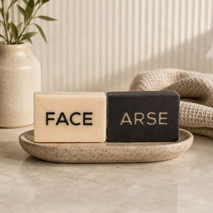 Two soap bars labeled 'FACE' and 'ARSE' on a stone tray with a plant and towel in the background.