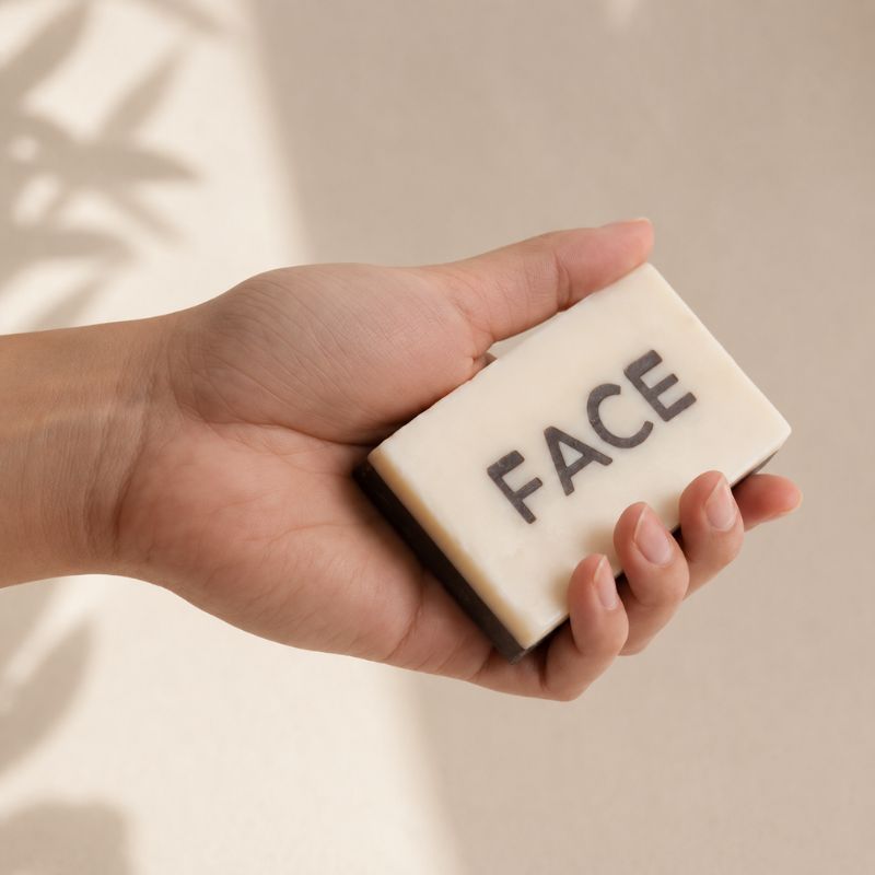 Hand holding a bar of soap with 'FACE' written on it against a neutral background