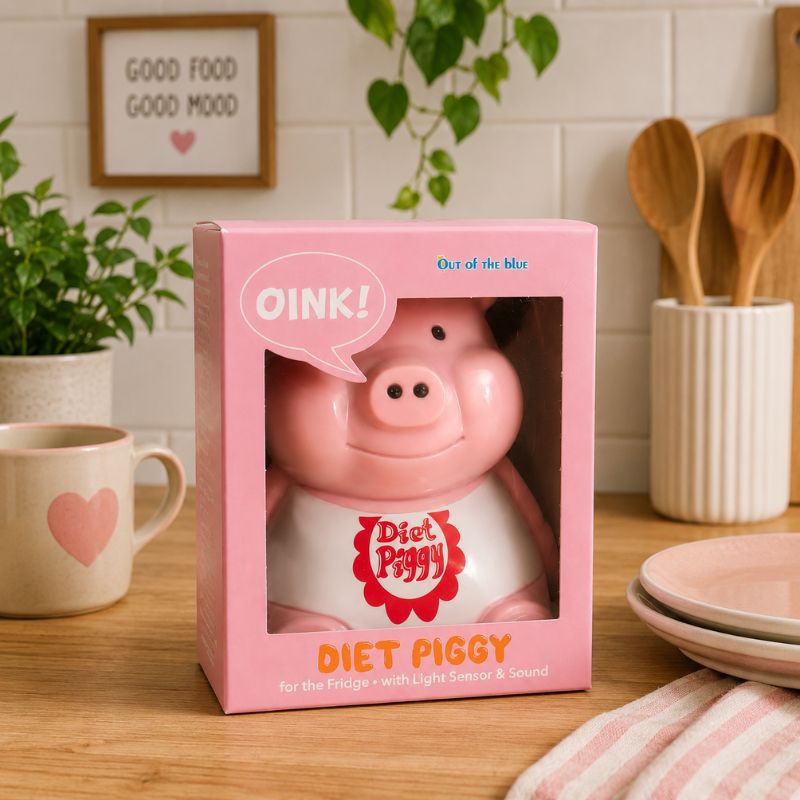 Pink 'Diet Piggy' toy in packaging on a kitchen counter with a mug and plants in the background.