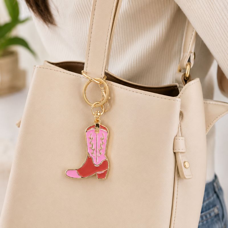 A close-up of a beige tote bag worn by a person, featuring a Pink Cowboy Boot Enamel Keyring with pink and red enamel and gold-tone metal.