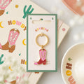 A western-themed Pink Cowboy Boot Enamel Keyring attached to a gold-tone ring and displayed on a green card. The card is placed on top of a matching printed card featuring cowboy boots and cacti, with a soft-focus bowl in the background.