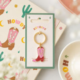 A western-themed Pink Cowboy Boot Enamel Keyring attached to a gold-tone ring and displayed on a green card. The card is placed on top of a matching printed card featuring cowboy boots and cacti, with a soft-focus bowl in the background.