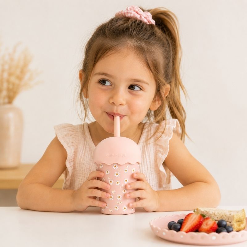 A young girl drinking from a Pink Daisy Mini Smoothie Cup & Straw 200 ml with a plate of fruit on a white surface.