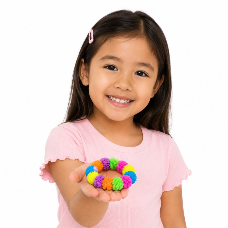 A young girl with long dark hair, wearing a pink shirt and holding a small hair bow made of colorful pom-poms against a plain white background.