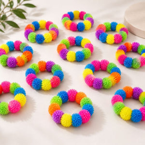 Multiple colorful Pom Pom Squishy Bracelet arranged on a light surface, with a small leaf in the top-left corner and part of a round object in the top-right. The rings are made of bright, fuzzy, spiky material in rainbow colors including pink, yellow, blue, green, orange, and purple.