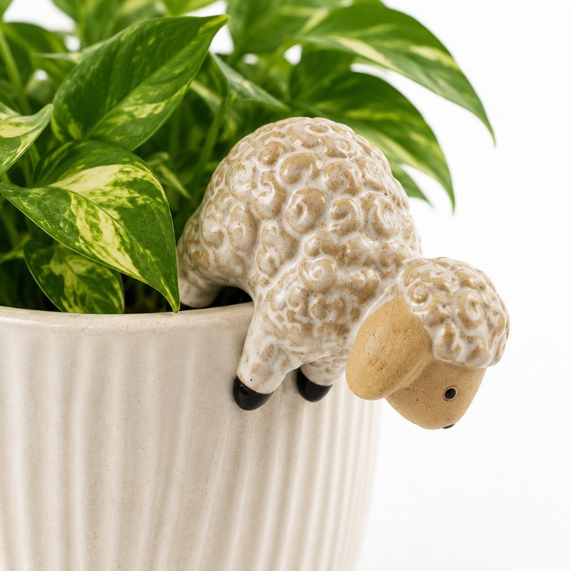 A ceramic sheep planter with a textured cream and tan body and a light tan face, shown overflowing green variegated pothos plant leaves from a ribbed white pot against a white background.-Sheep Pot Hanger Ceramic