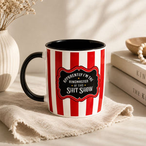 A ceramic mug with red and white vertical stripes and a black handle, featuring a black label with white text that reads, 'APPRORENTLY I'M THE RINGMASTER OF THIS SHIT SHOW.' It is styled on a beige cloth with books, a wooden bowl, and dried flowers in the background.