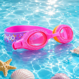 Rock Candy Pink Swim Goggles displayed beside a sparkling swimming pool with soft water reflections, highlighting the vibrant pink color and glittering details.