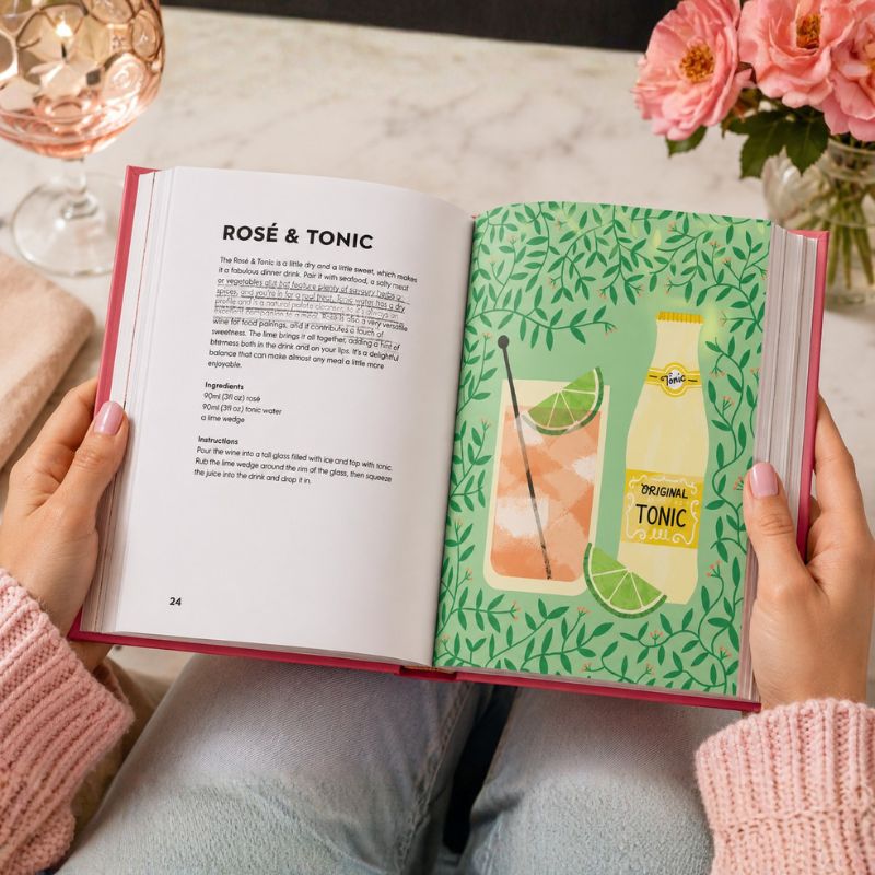 Open book with a Rosé & Tonic recipe and a lime illustration on a table with flowers and a glass.