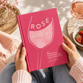 Person holding a pink book titled 'Rosé Made Me Do It' with a glass of rosé and strawberries in the background.