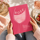 Person holding a pink book titled 'Rosé Made Me Do It' with a glass of rosé and strawberries in the background.