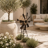 Metal garden windmill stake displayed on a stylish outdoor patio with beige seating, potted olive trees, and white flowers.