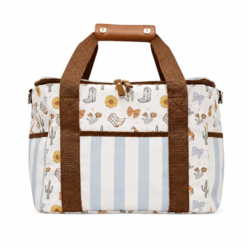 A Saddle & Bloom Cooler Bag with a western-themed print featuring cowboy boots, cacti, and sunflowers, accented by brown canvas handles and trim and light blue vertical stripes on the front panel.