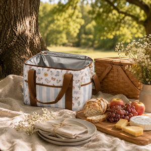 A Saddle & Bloom Cooler Bag with brown handles sits on a beige blanket outdoors beside a picnic basket, bread, grapes, cheese, and flowers under a tree.