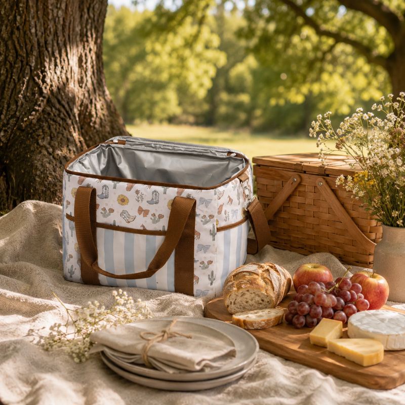 A Saddle & Bloom Cooler Bag with brown handles sits on a beige blanket outdoors beside a picnic basket, bread, grapes, cheese, and flowers under a tree.