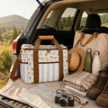 A Saddle & Bloom Cooler Bag with brown handles and a striped base is displayed in the open trunk of a car, surrounded by camping accessories including a canvas tote bag, a straw hat, water bottles, folded blankets, hiking boots, and a camera, with a scenic outdoor landscape in the background.