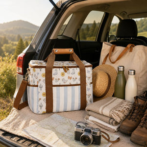 A Saddle & Bloom Cooler Bag with brown handles and a striped base is displayed in the open trunk of a car, surrounded by camping accessories including a canvas tote bag, a straw hat, water bottles, folded blankets, hiking boots, and a camera, with a scenic outdoor landscape in the background.