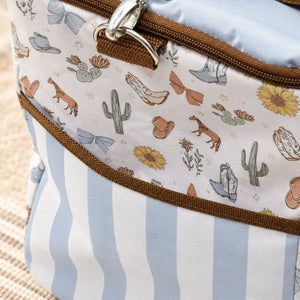 Close-up of a Saddle & Bloom cooler bag with a western-themed print on the upper section and light blue and white vertical stripes on the lower section. The bag features brown trim, a silver zipper pull, and a light blue fabric lining.