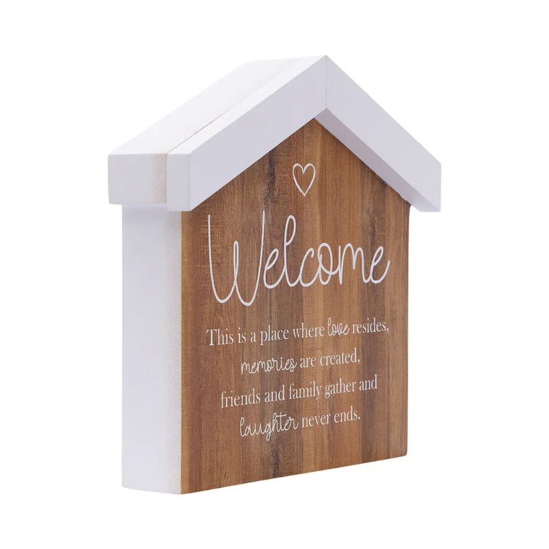 A house-shaped wooden wall decor sign with a white painted frame and a natural wood front panel. The panel features the word 'Welcome' in white script, a small heart outline, and a sentimental quote below, all set against a plain white background.