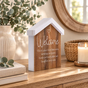 A Saddle & Bloom Welcome Blockword is styled on a wooden table with a candle, woven basket, books, and greenery in a softly lit interior setting.