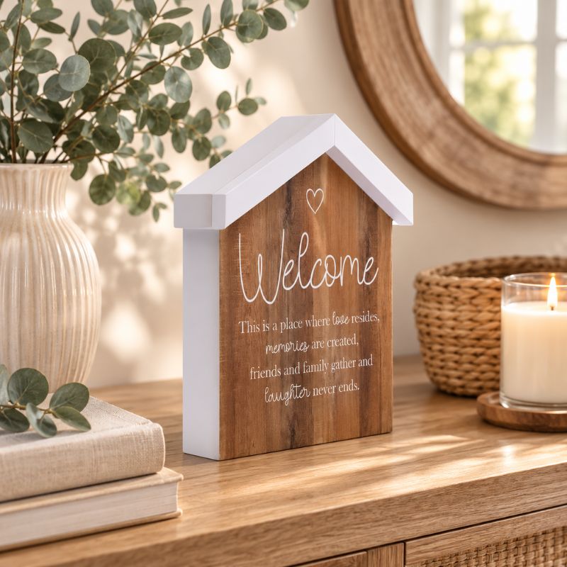 A Saddle & Bloom Welcome Blockword is styled on a wooden table with a candle, woven basket, books, and greenery in a softly lit interior setting.