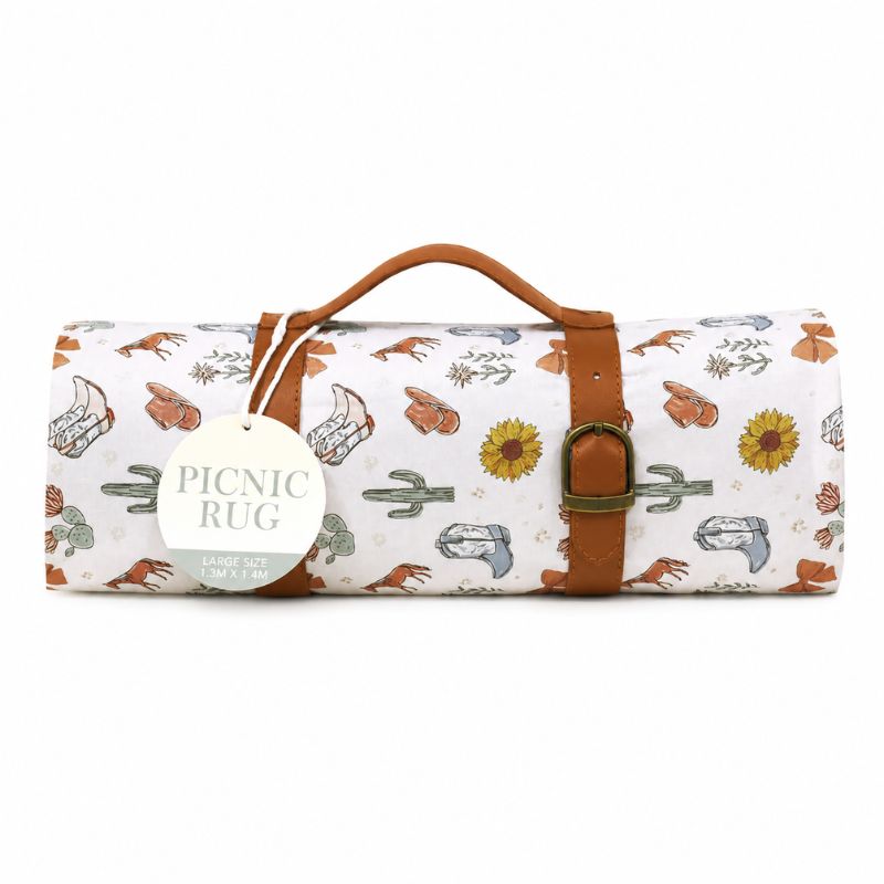A Saddle & Bloom Picnic Rug with a white background and a western-themed print featuring boots, cacti, sunflowers, and rattlesnakes. It has a brown leather handle and strap with a brass buckle, and a round white tag that reads 'PICNIC RUG'.