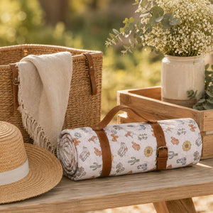 A Saddle & Bloom Picnic Rug, styled on a wooden bench next to a straw hat, a woven basket, and a floral arrangement.