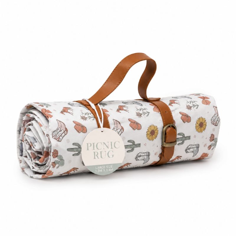 A rolled-up Saddle & Bloom Picnic Rug with a brown leather carry handle and strap, featuring a western-themed print of boots, cacti, and sunflowers on a white background. A paper tag reads 'PICNIC RUG'.