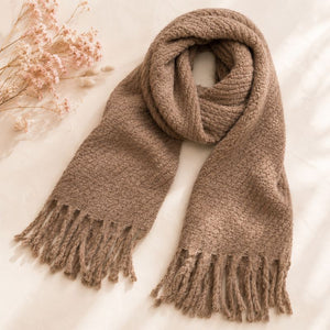 A Sand Chunky Knit Winter Scarf with Fringe, styled in a loop with fringed ends on a soft cream fabric background with dried flowers in the corner.