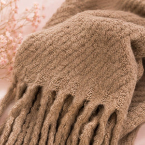 A close-up of a Sand Chunky Knit Winter Scarf featuring knotted fringe details and draped over a soft pink background with dried floral accents.