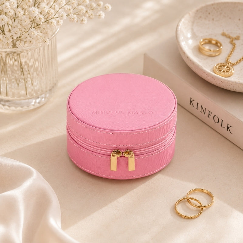 A Small Jewellery Case – Berry with a gold zipper and dual pull tabs, styled on a beige surface with decorative props including rings, a vase of flowers, and a book.