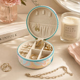 A Small Jewellery Case – Haven with a pastel multicolor floral pattern and turquoise trim, displayed on a tabletop with rings, earrings, and a chain around it. A candle, books, and flowers are visible in the styled setting.