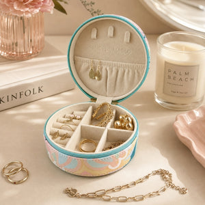 A Small Jewellery Case – Haven with a pastel multicolor floral pattern and turquoise trim, displayed on a tabletop with rings, earrings, and a chain around it. A candle, books, and flowers are visible in the styled setting.