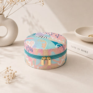 A small jewelry case – Haven with a colorful abstract print in pink, blue, and beige, featuring a teal zipper and gold-tone dual pull hardware. It is styled on a light beige surface next to a white ceramic vase, a book, and a small dish.