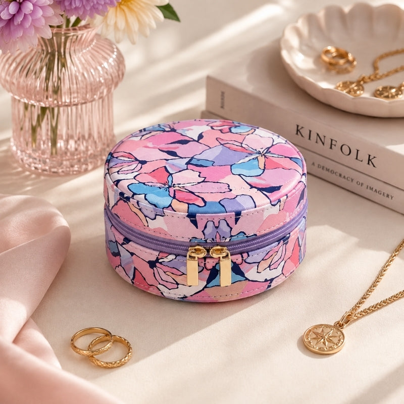 A Small Jewellery Case – Willow with a pink, purple, and blue floral pattern sits on a beige surface surrounded by gold jewelry, a pink glass vase with flowers, and a book.