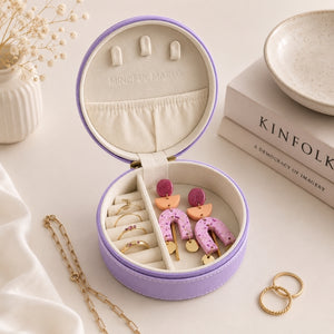 A small travel jewelry case, violet, is shown open, filled with earrings and rings, styled on a light beige surface with books, a ceramic dish, and a small vase in the background.