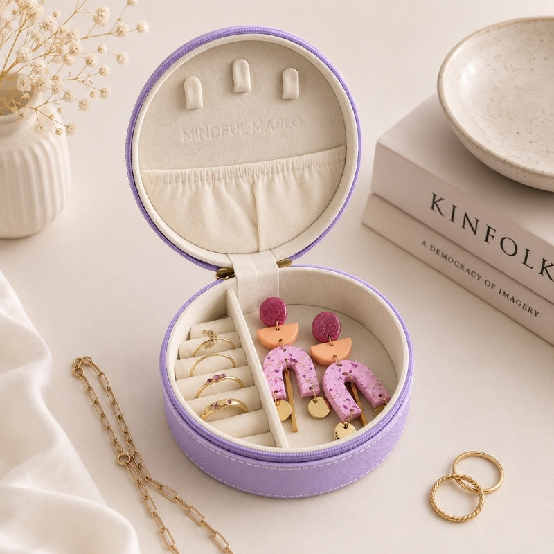 A small travel jewelry case, violet, is shown open, filled with earrings and rings, styled on a light beige surface with books, a ceramic dish, and a small vase in the background.