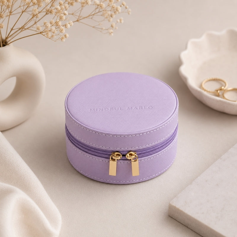 A Small Travel Jewellery Case Violet with a gold zipper and gold pull tabs, featuring the debossed brand name 'MINIDFUL MARLO' on the lid. The box is styled on a neutral beige surface with decorative props including a ceramic vase with dried flowers, a white dish with gold rings, and a marble block.