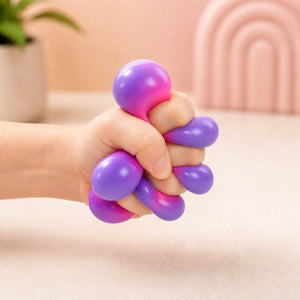 A hand squeezing a purple and pink stress ball toy with multiple finger-like loops, shown against a soft beige indoor background with a blurred plant and arch decor.