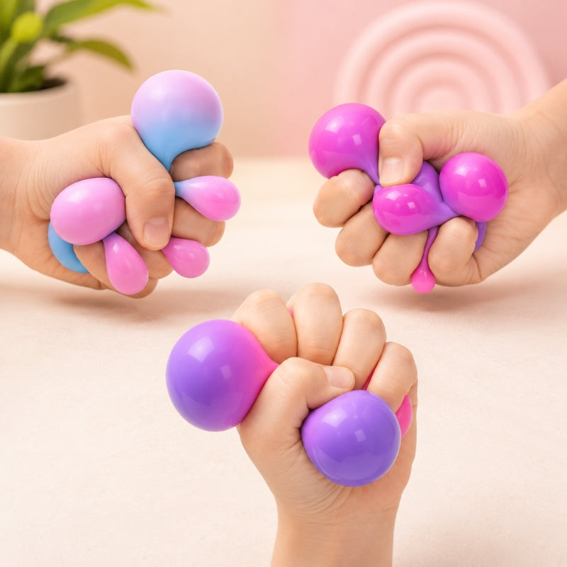 Three hands squeezing colorful stress balls in a pink studio setting. The stress toys are glossy and come in pastel gradient colors including pink, purple, and blue.