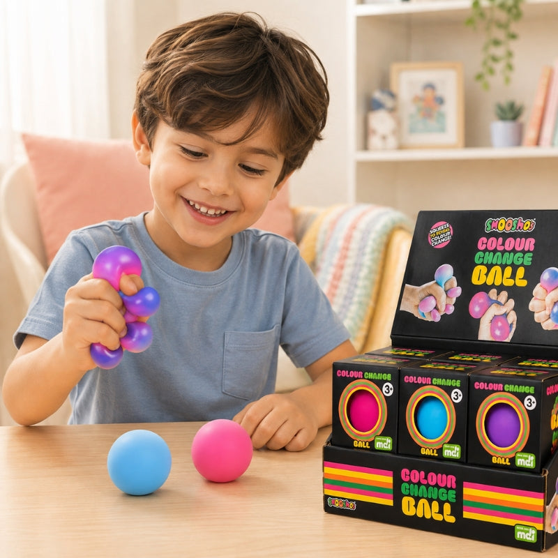 A young boy smiling and holding a purple and blue squeeze ball toy at a wooden table, with two loose balls in front of him and a retail display box of 'Squishies Colour Change Ball' toys to his right.