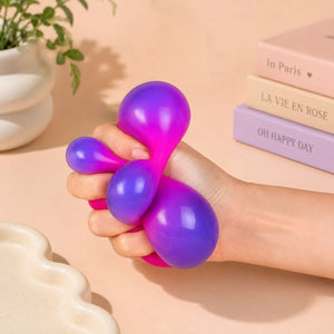 A hand squeezing a Smoosho's Colour Change Cube – Sensory Squishy Toy in purple and hot pink, set against a warm beige background with decorative books and a plant.