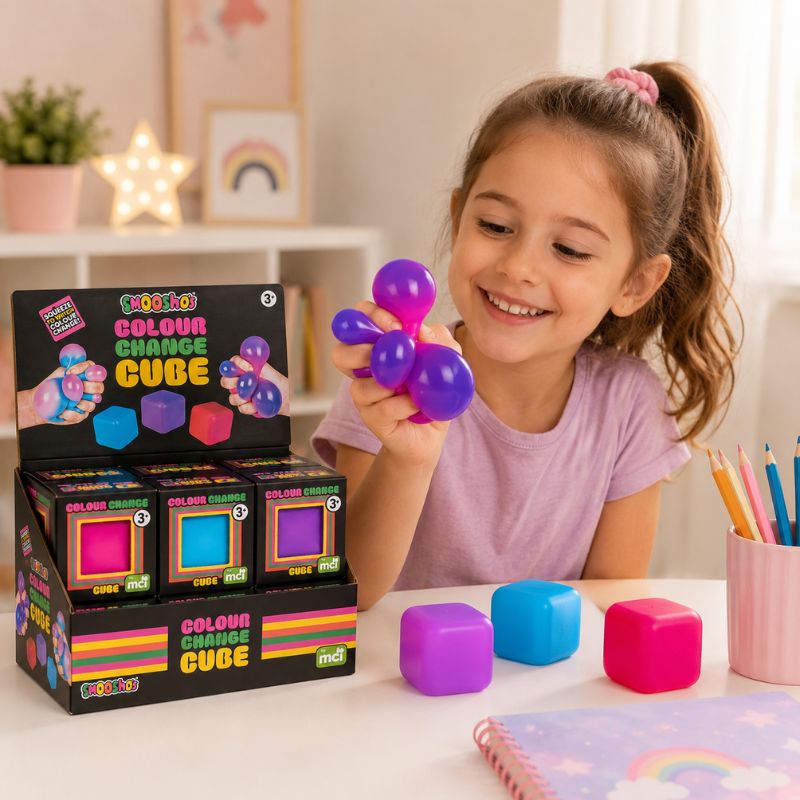 A young girl with a ponytail smiles while holding a Smoosho's Colour Change Cube – Sensory Squishy Toy. In front of her is a retail display box of 'Color Change Cube' toys by 'mii', along with several colorful squeeze cubes on a white table in a brightly lit, child-friendly room.