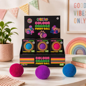 A set of three furry ball foot massagers in bright pink, purple, and blue, displayed in front of their black retail packaging on a tabletop. The background is a warm, decorated room with a potted plant, bunting, a rainbow, and a framed sign.