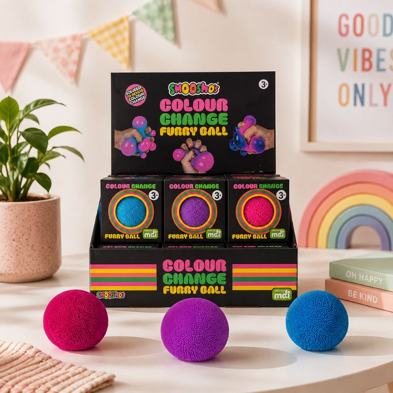 A set of three furry ball foot massagers in bright pink, purple, and blue, displayed in front of their black retail packaging on a tabletop. The background is a warm, decorated room with a potted plant, bunting, a rainbow, and a framed sign.