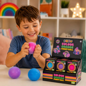 A smiling young boy in a blue t-shirt holds a pink spiky squishy ball, with two more balls in purple and blue on the table next to a black display box of 'Goo goo Colour Change Furry Ball' toys. The background shows a softly blurred, warm-toned children's room shelf.