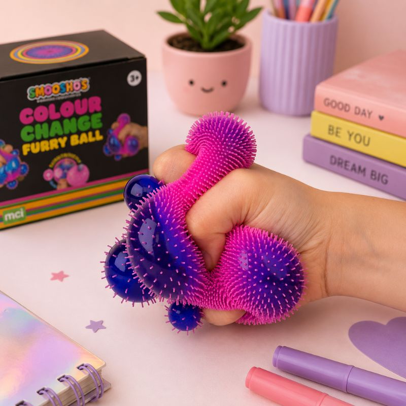Smoosho’s Colour Change Furry Ball being squeezed in a hand on a pastel desk, with the product box, plant, books, and stationery in the background.