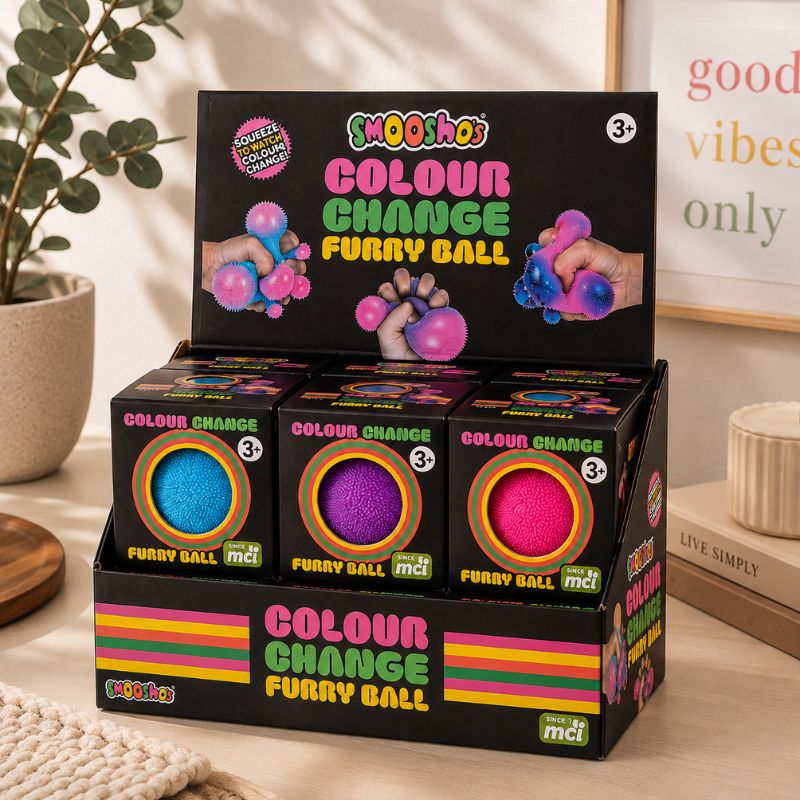A retail display box of mcti 'Shoo Shoo' Colour Change Furry Ball toys, showing three colorful balls (blue, purple, and pink) inside black packaging with retro-style pink, yellow, and green lettering, set on a wooden table next to a potted plant.