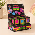 A retail display box filled with SmooSho Colour Change Furry Cube toys, shown on a warm beige tabletop with a potted plant and pastel rainbow decor in the background.